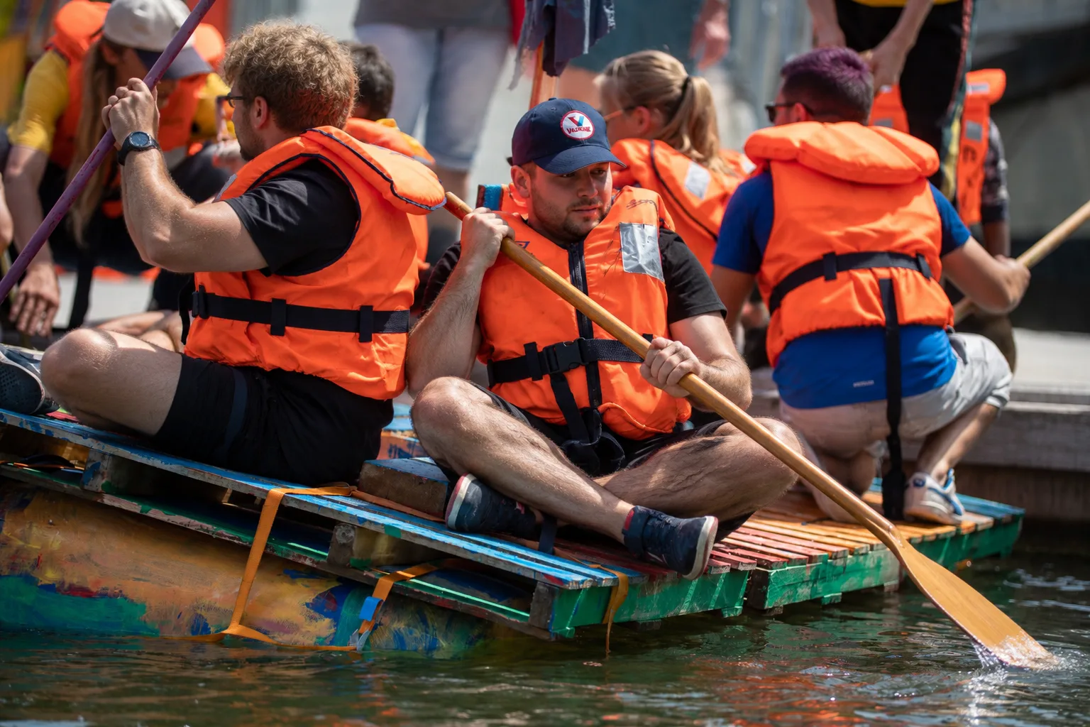 Two participants in orange life vests paddle a colourful pallet raft.