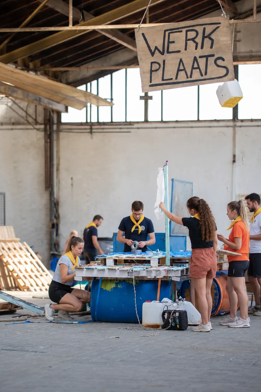 Participants build their raft in a workshop, beneath a sign reading 'Werkplaats'.