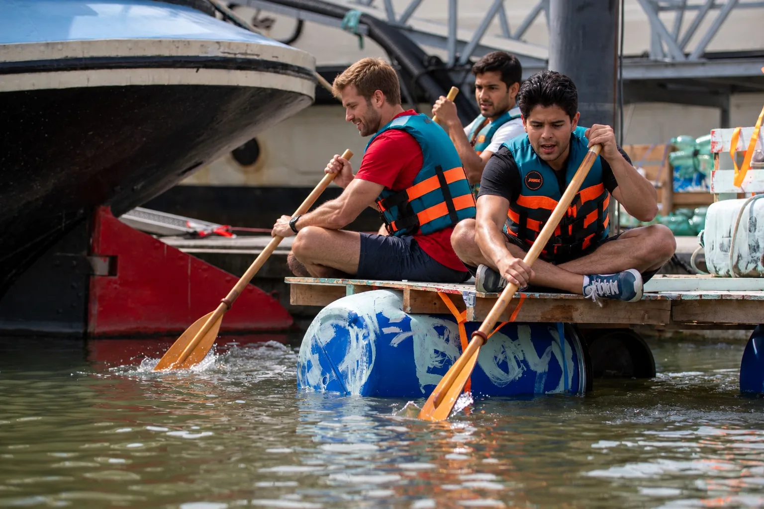 Three participants paddle hard on a raft built on blue barrels, past a boat.